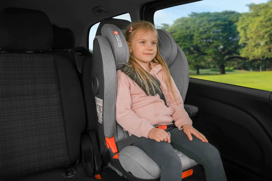 A young girl in a pink jacket is seated comfortably in a gray high back booster car seat by Zopa, featuring vibrant orange accents and secure Isofix attachments, in the back seat of a car with a scenic park outside.