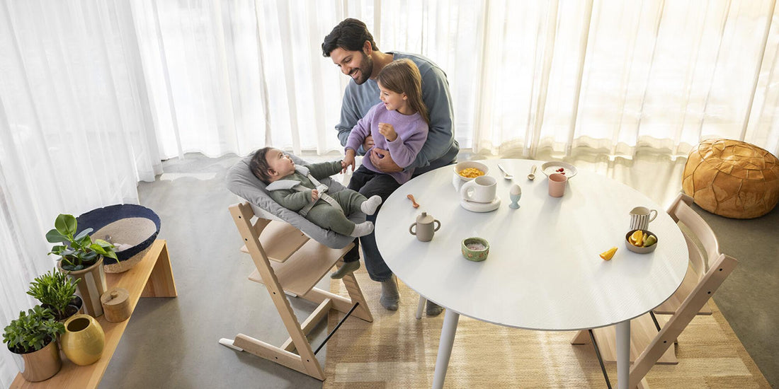 A father smiles at his two kids in a sunny dining room. A baby sits in a Stokke Tripp Trapp chair, while the older child in purple is in his arms, with breakfast on the table.