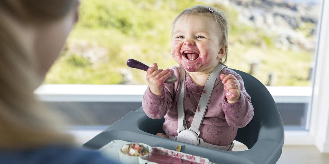 A joyful baby, covered in berry puree, laughs heartily while sitting in a Stokke Clikk high chair. Holding a purple spoon and wearing a bib, the moment captures the messy delight of a child's mealtime.