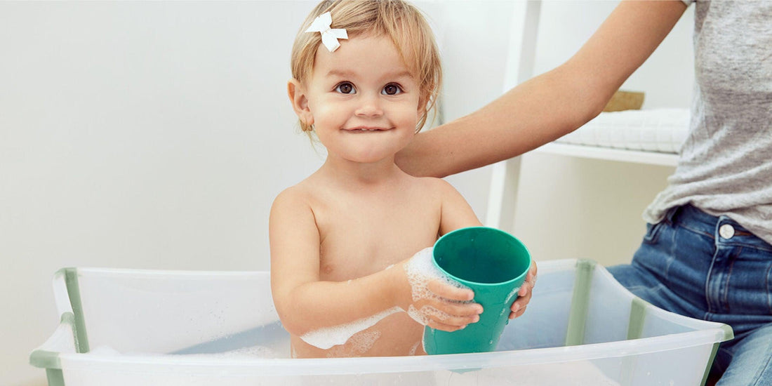 A smiling toddler with a playful bow in her hair enjoys bath time in a Stokke Flexi Bath, holding a green cup with soapy water, with a parent's arm gently supporting her.