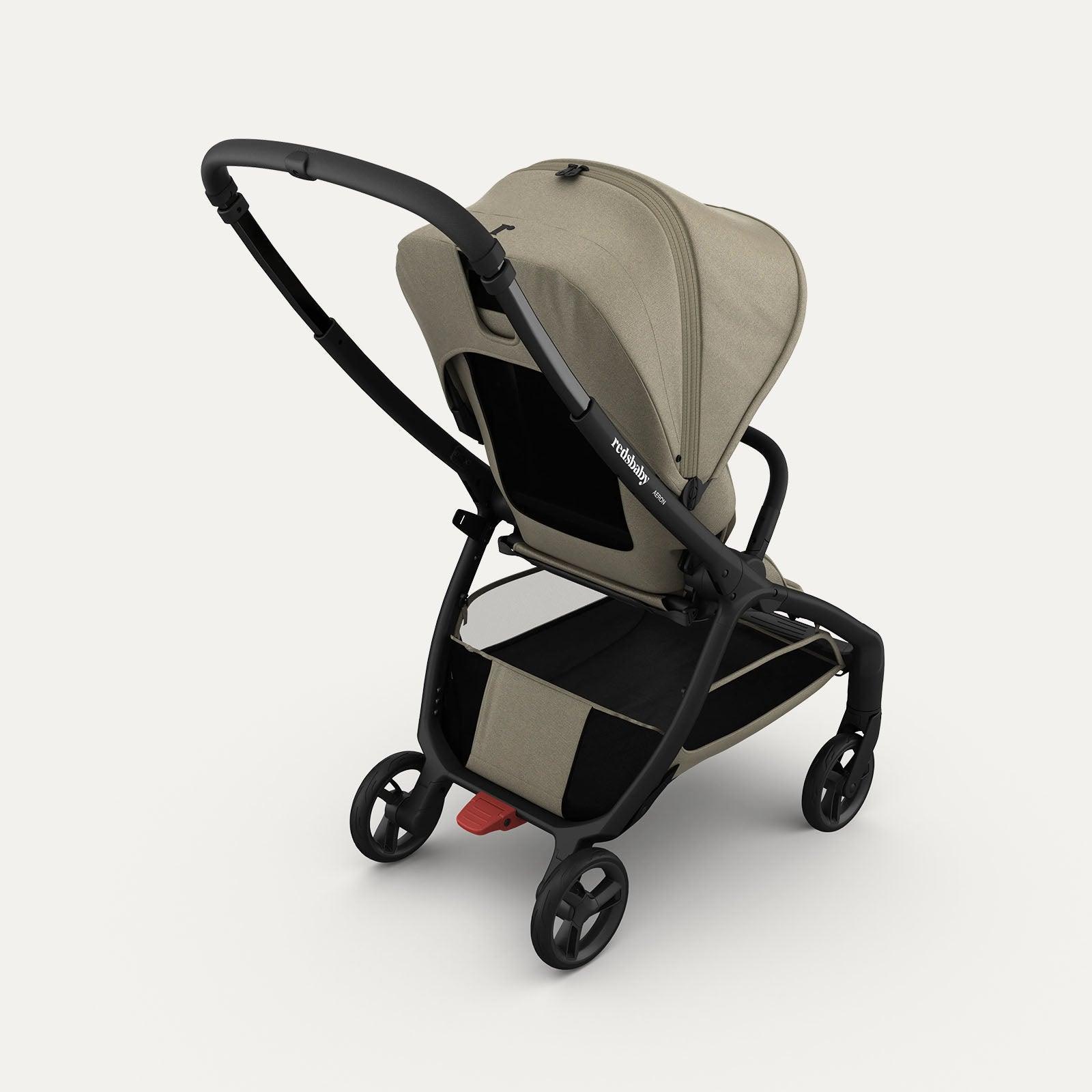 Rear view of the taupe-colored Redsbaby Aeron stroller with a black frame. The stroller features a large canopy, mesh back panel for ventilation, spacious storage basket underneath, and no-puncture wheels with a red brake pedal on the rear wheels. The sleek and modern design highlights its practicality and functionality. Rear view of the taupe-colored Redsbaby Aeron stroller with a black frame. The stroller features a large canopy, mesh back panel for ventilation, spacious storage basket underneath, and no-puncture wheels with a red brake pedal on the rear wheels. The sleek and modern design highlights its practicality and functionality.