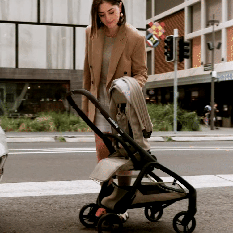 A woman in a beige blazer and white skirt is folding a taupe-colored Redsbaby Aeron stroller with a black frame on a city street. The background features urban buildings, traffic lights, and greenery, showcasing the stroller's ease of use and portability in an urban environment. A woman in a beige blazer and white skirt is folding a taupe-colored Redsbaby Aeron stroller with a black frame on a city street. The background features urban buildings, traffic lights, and greenery, showcasing the stroller's ease of use and portability in an urban environment.