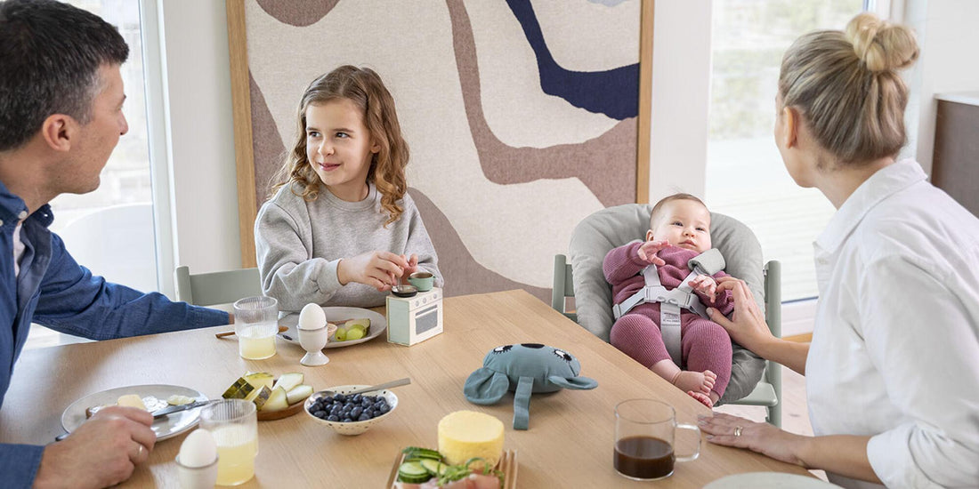 A family enjoys a meal together; a father to the left looks at his daughter who's smiling and playing with a toy radio. On the right, a mother lovingly gazes at her baby secured in a Stokke Tripp Trapp with a newborn bouncer set.