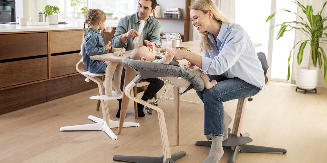 A family shares a meal, with each member seated on a Stokke Nomi chair, showcasing its versatility. The mother engages with the baby in a bouncer attachment, while the father feeds the older child, creating a cozy, connected atmosphere.