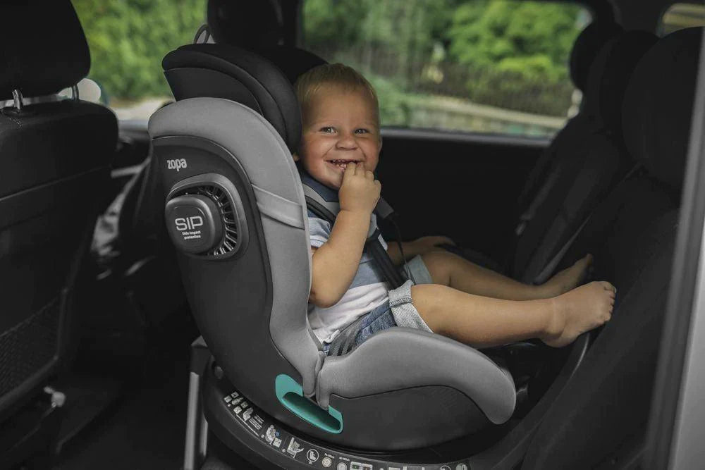 A cheerful toddler sits comfortably in a Zopa Voyager Max car seat, with his finger on his lips as if pondering, the seat's grey and black design and side impact protection are visible.