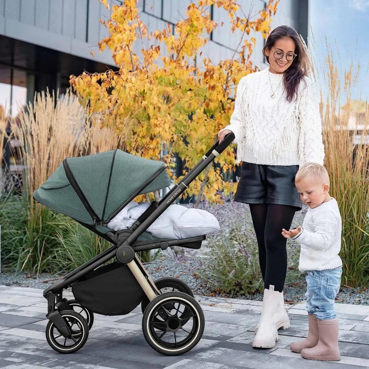 A stylish young mother stands with a Zopa Move Cross 2 stroller in parent-facing position, showcasing its luxurious Antique Green color with gold accents. She's dressed in a chic white cable knit sweater, black leather shorts, and white ankle boots, accessorized with glasses and a necklace. Next to her, a toddler in a white sweater and blue jeans looks curiously at something in his hand. They are outdoors, with vibrant autumn leaves in the background, creating a warm and fashionable family scene.