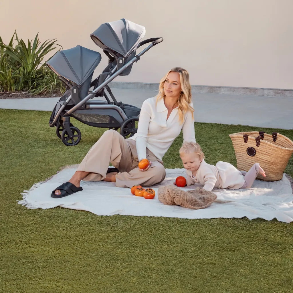 A relaxed outdoor setting features a woman and a toddler sitting on a white blanket on the grass, with a Redsbaby NUVO stroller in twin/tandem mode nearby. The stroller is sleek with two slate blue seats facing each other, suitable for twins or siblings of different ages. The woman, dressed in a cream sweater and taupe trousers, interacts with the child who is playing with some fruit. A straw basket adds a picnic-like atmosphere to this serene family moment.