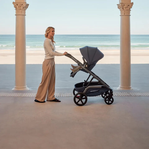 A woman stands on a covered patio by the beach, gently pushing a Redsbaby NUVO stroller. She is dressed in a casual cream sweater and taupe trousers, complementing the stroller's modern design in a matching slate blue color. The calm ocean and clear sky create a serene backdrop, while the stroller's design, featuring a large canopy and streamlined frame, is highlighted by the natural daylight.