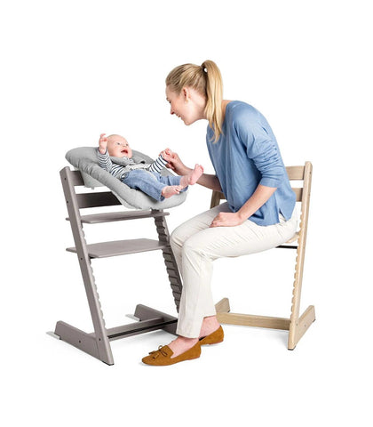 A mother sitting on a Stokke Tripp Trapp chair, while playing with her baby also in a Stokke Tripp Trapp chair but with a newborn set on top instead.