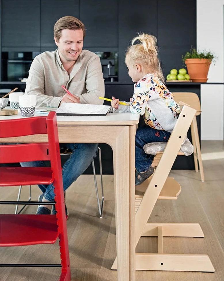A father and his daughter sitting in a kitchen with the child sitting on a Stokke Tripp Trapp chair in natural colour. A red Tripp Trapp chair can also be seen in the side.