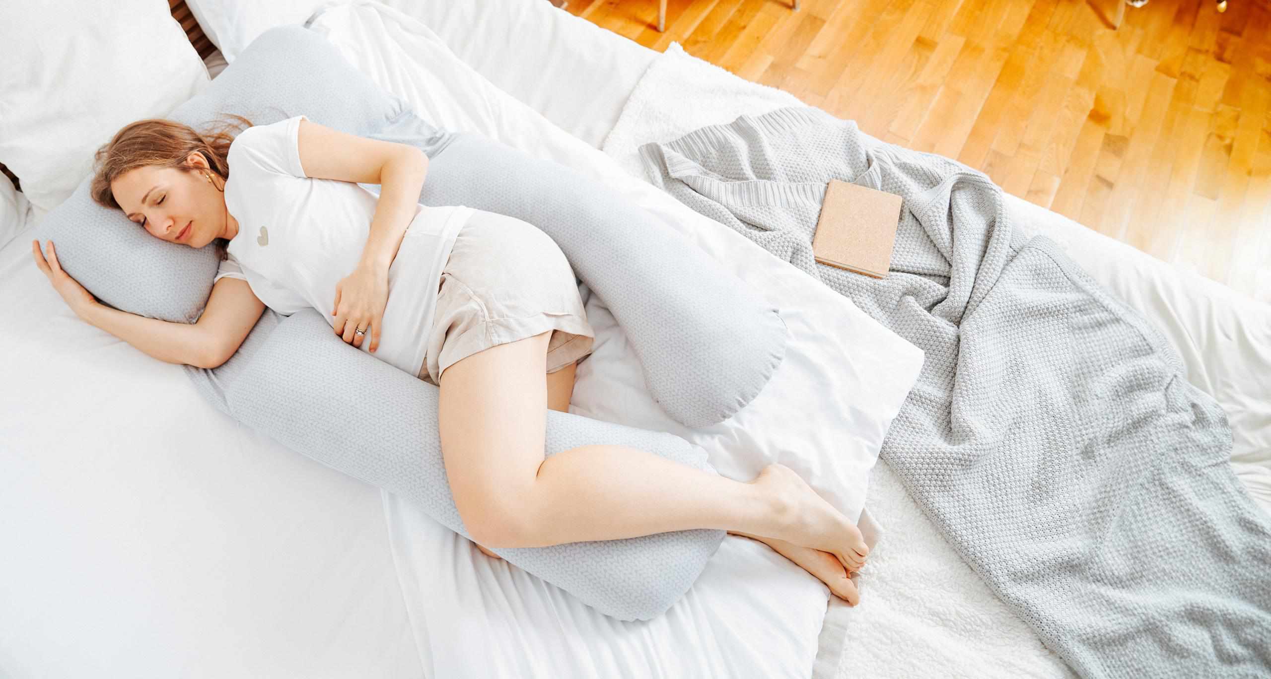 A peaceful image of a pregnant woman resting comfortably on a large Babymoov pregnancy pillow in mineral grey. The pillow's extra-long body contours around her, supporting her belly and legs as she sleeps on her side. She is dressed in light casual clothes and is positioned on a white bed with crisp linens. A knitted grey throw and an open book lie beside her, suggesting a relaxed and cozy atmosphere. The room is bright with natural light reflecting off the wooden floor, adding warmth to the serene scene.